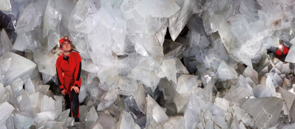 Person in a red protective suit and helmet standing among jagged white crystals in a cavern